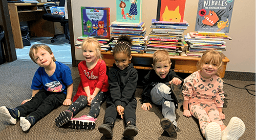 Five young children sit on the floor in front of stacks of children's books and colorful book covers, smiling at the camera.