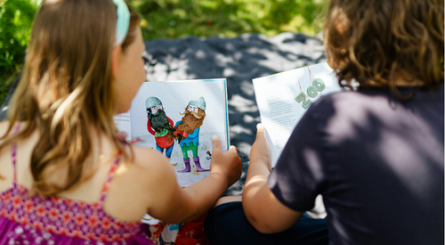 Five young children sit on the floor in front of stacks of children's books and colorful book covers, smiling at the camera.
